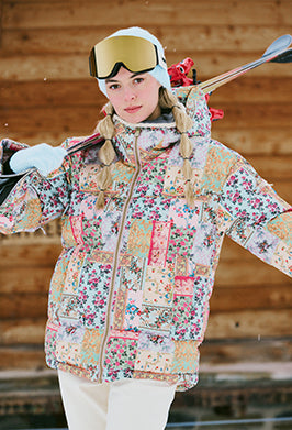 A woman in a floral patchwork winter jacket and white ski pants stands outdoors holding skis. She wears goggles, white gloves, and a beanie, with her blonde hair in braids. Snow falls gently and a wooden wall is behind her.