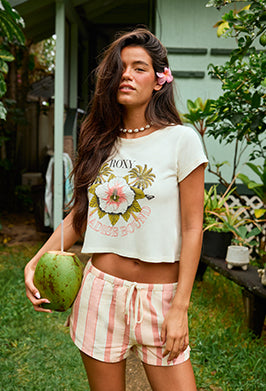 Woman holding a coconut in a tropical setting wearing a floral crop top and checkered shorts.