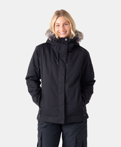 A person with blonde hair smiles, wearing a black winter jacket with a fur-lined hood, standing with hands in pockets against a plain light background.