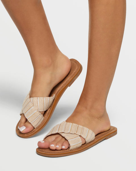 A person wears Roxy Andreya Sandals—tan woven slides with crisscross straps and brown soles—on a white background. Their toenails are painted white.