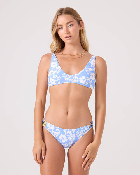A woman with long blonde hair and minimal jewelry wears the Roxy Golden Glow Reversible Scoop Bikini Top featuring light blue, white floral patterns, and gold accents. She stands against a plain background with a relaxed expression.
