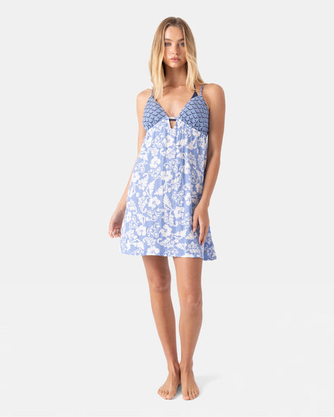 A woman stands barefoot against a plain background, wearing the Roxy Golden Glow Cover Up Dress—a light blue and white floral sundress with a fitted, patterned bodice and thin straps, perfect as a strappy dress or stylish beach cover-up.