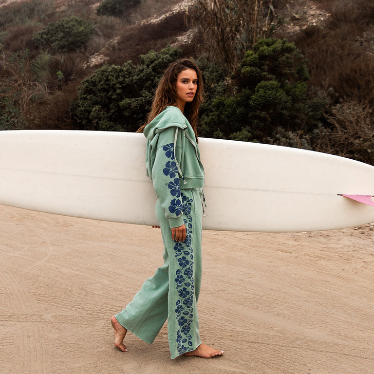A woman in a light green floral tracksuit carries a white surfboard under her arm while walking barefoot on a sandy path, with greenery and hills in the background.