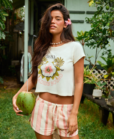A woman with long brown hair and a pink flower in her hair stands outside, holding a coconut. She wears a white floral crop top that says Roxy Paradise Bound and pink-and-white striped shorts. Green plants and a house are in the background.