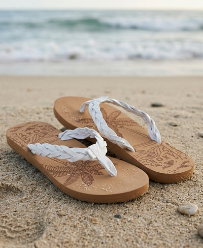 A pair of tan flip-flops with white braided straps rests on sandy beach near the shoreline, with gentle ocean waves in the background.