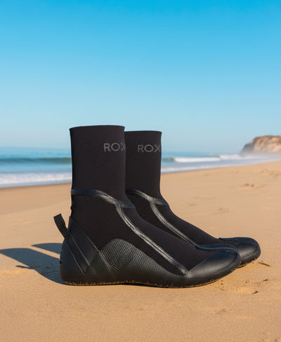 A pair of black ROXY surf booties stand upright on sandy beach with gentle waves and cliffs in the background under a clear blue sky.