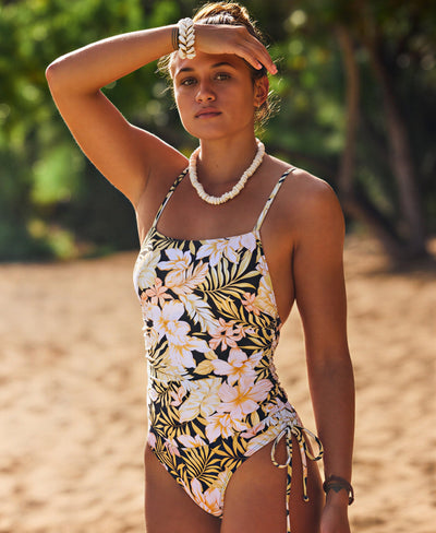 A person wearing a floral one-piece swimsuit, shell necklace, and bracelet stands on a sandy beach with one arm raised to their forehead. Lush greenery is blurred in the background.