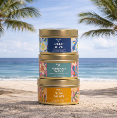 Three colorful tins of Roxy candles labeled Deep Dive, Hibiscus Wave, and Sun Drift are stacked on a sandy beach, with palm trees and the ocean in the background under a partly cloudy sky.