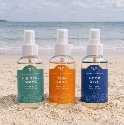 Three bottles of Roxy Body Mist—Hibiscus Wave, Sun Drift, and Deep Dive—are arranged on sandy beach with the ocean and sky in the background.