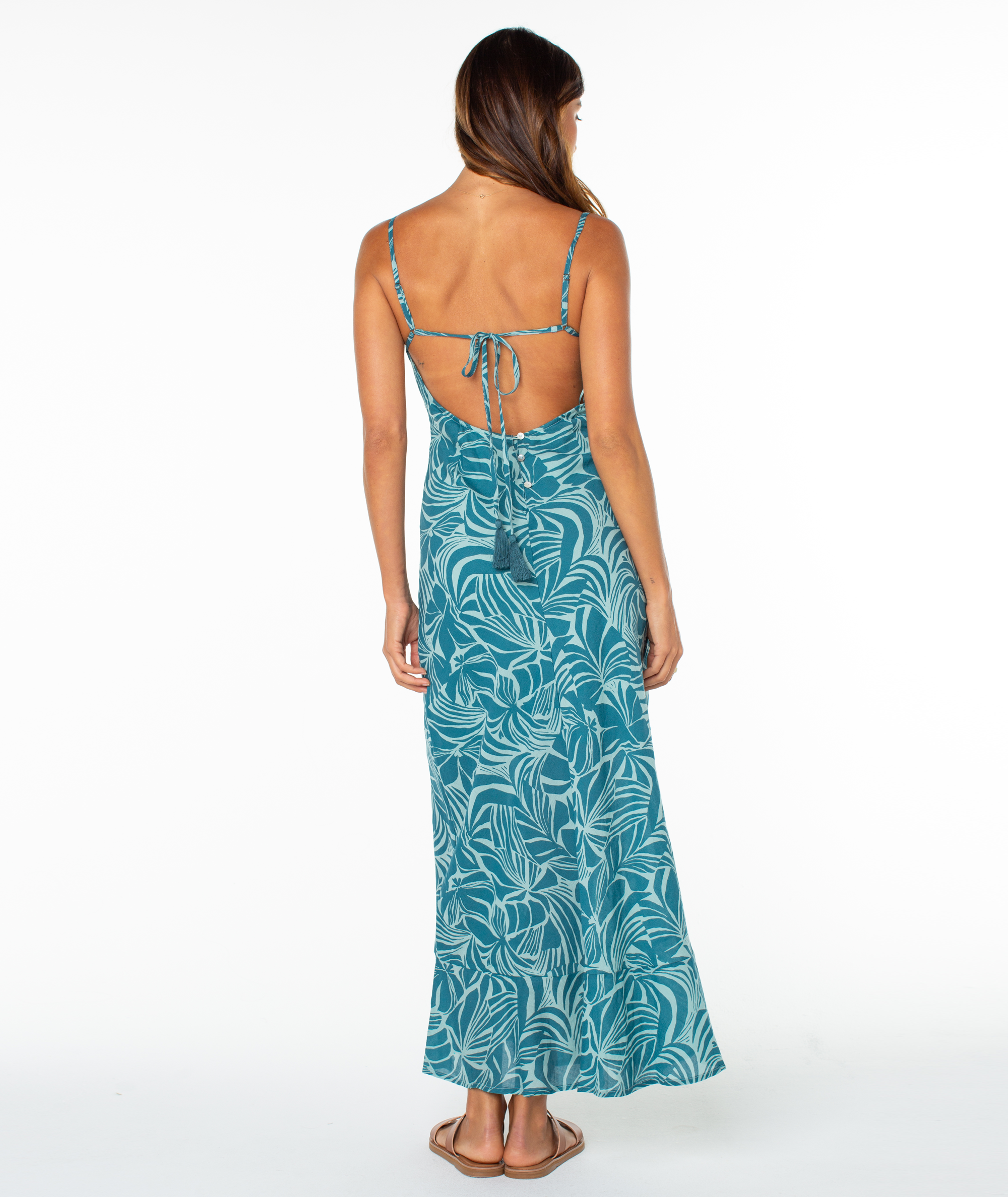 A woman with long brown hair wears the Roxy®一分钟极速赛车168开奖结果记录体彩历史号码查询 Lost Paradise Dress, a teal maxi dress with a white leaf print and an open, strappy back. She pairs it with sandals and stands on a white background, facing away from the camera.