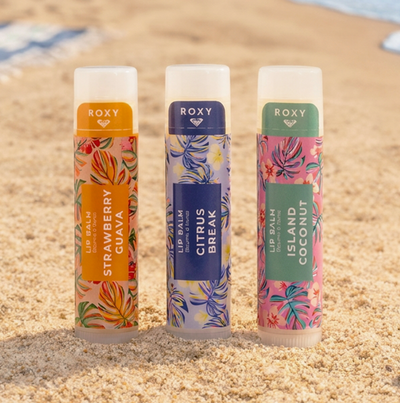 Three colorful Roxy lip balm tubes—Strawberry Guava, Citrus Break, and Island Coconut—stand upright on sandy beach with ocean water in the background.