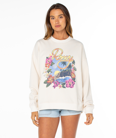 A woman wears the Roxy Lineup Oversized Crew, a cream-colored sweatshirt made from a soft cotton blend with a sun-washed finish and colorful floral, mountain, and sunset graphics, paired with light denim shorts against a plain white background.