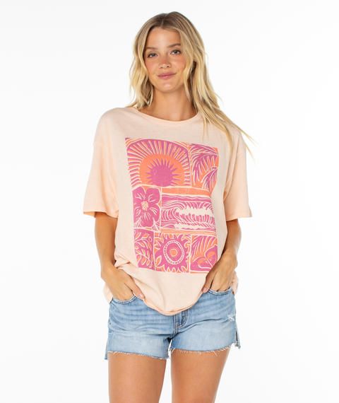 Wearing the Roxy Beach Things Tee, a woman in denim shorts smiles at the camera. The light pink oversized tee features a bold pink and orange floral design, set against a white background.