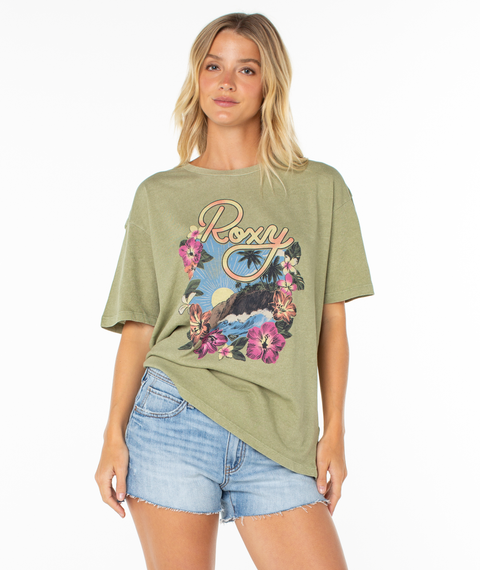 A woman stands against a white background wearing the Roxy Surfin Tee, a green graphic top with tropical floral and surfboard designs, relaxed drop shoulders, and light denim shorts.