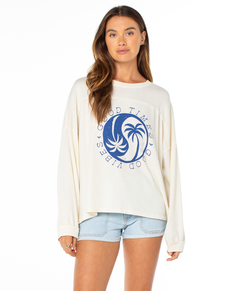 A woman stands against a plain white background wearing Roxy's Changing Tides Top, a cream slouch pullover with a blue circular Good Times Good Vibes palm tree graphic, paired with light blue denim shorts—ideal for spring.